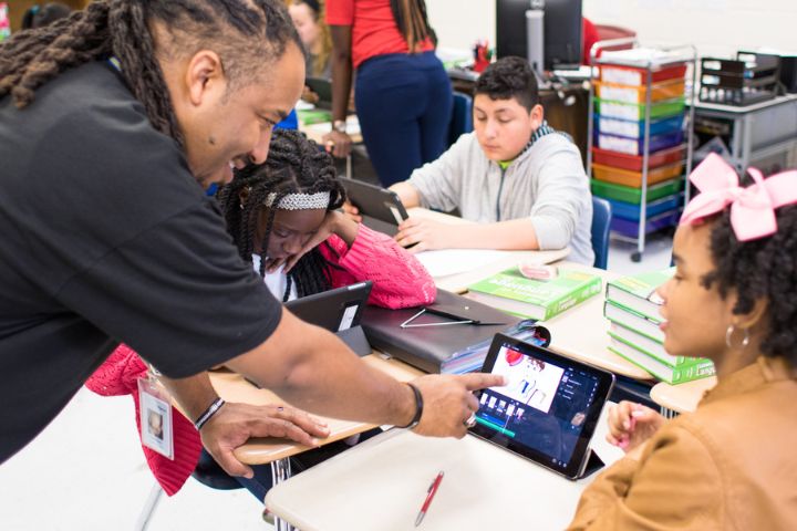 A teacher points to a tablet screen, assisting three students seated at a classroom table with their digital assignments.
