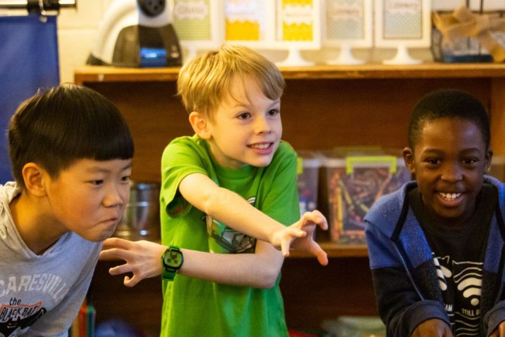 Three young boys in casual clothes are standing indoors, playfully posing with their arms outstretched and smiling.