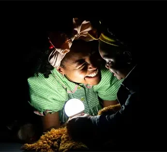 Two people, one dressed in a green costume with bows and the other in dark clothes, smile at each other while illuminated by a small glowing light in a dark setting.