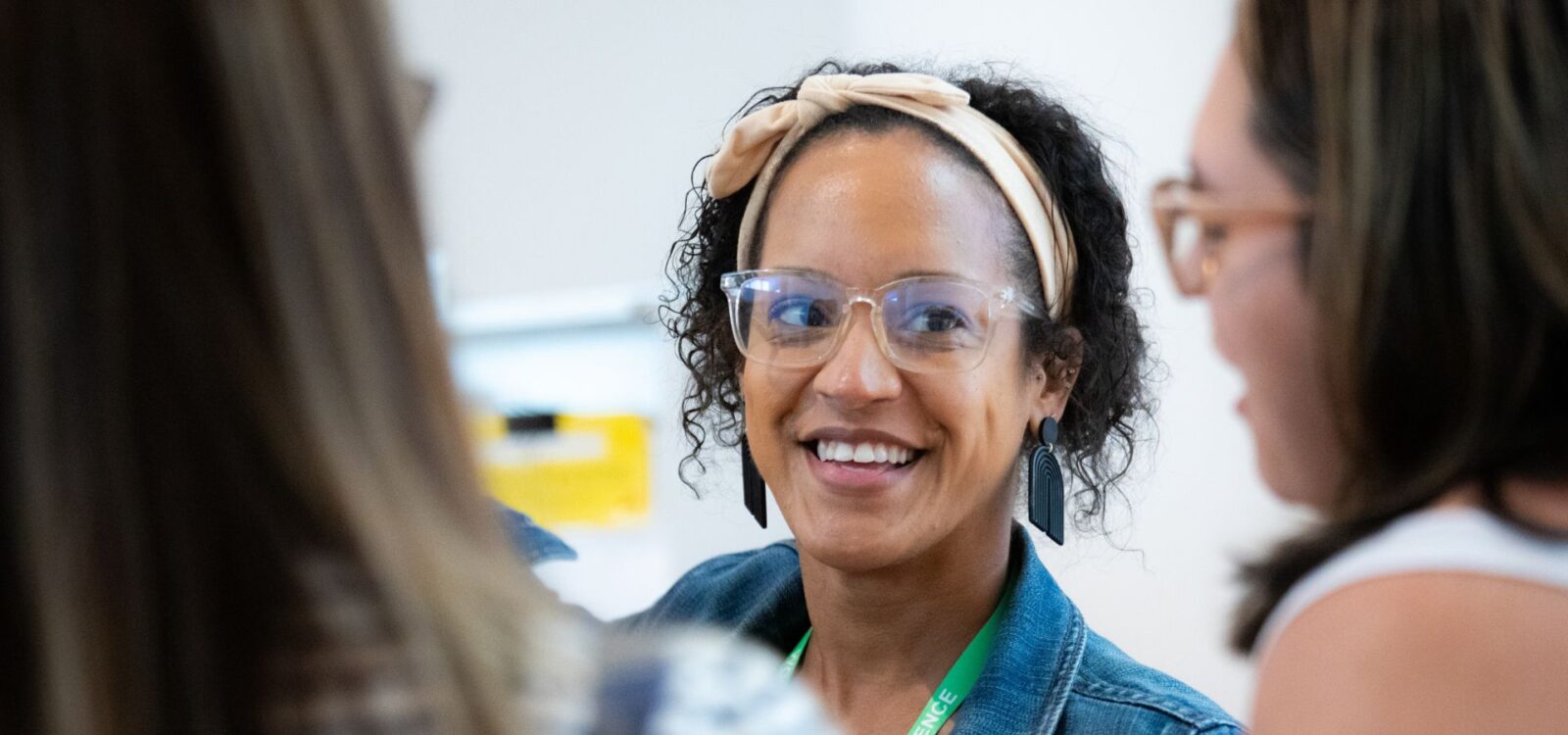 A woman wearing glasses and a headband smiles while talking with two other people in a bright indoor setting.