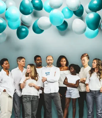 A group of nine people in casual white clothing stand together under teal and white balloons against a light blue background.