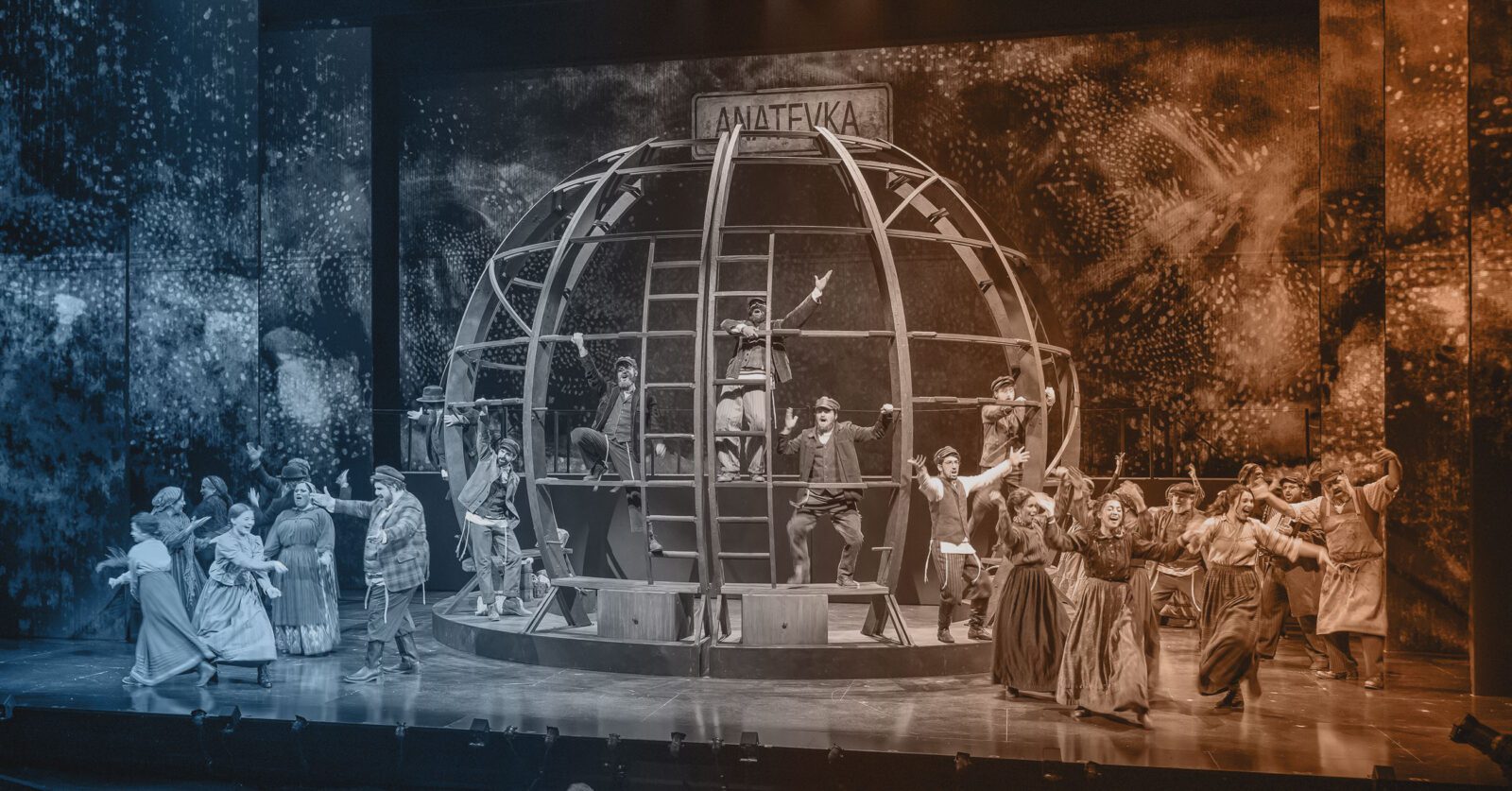 A theatrical performance shows actors dancing and interacting around a large spherical set piece with a sign reading “Anatevka” on stage.