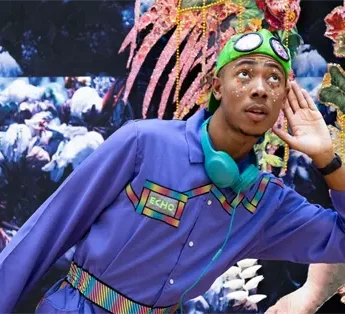 A person wearing a blue jumpsuit, green goggles, and teal headphones poses with one hand to their ear in front of a colorful, textured backdrop.