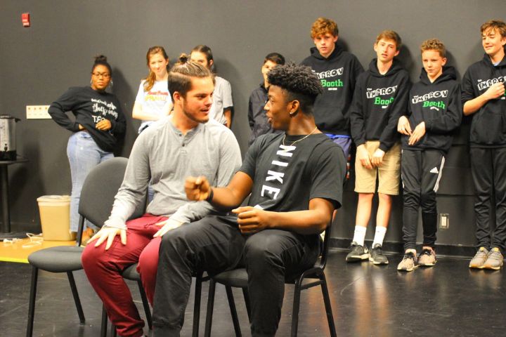 Two young men sit and talk in the foreground, while a group of students stand and watch in the background in a classroom or rehearsal space.