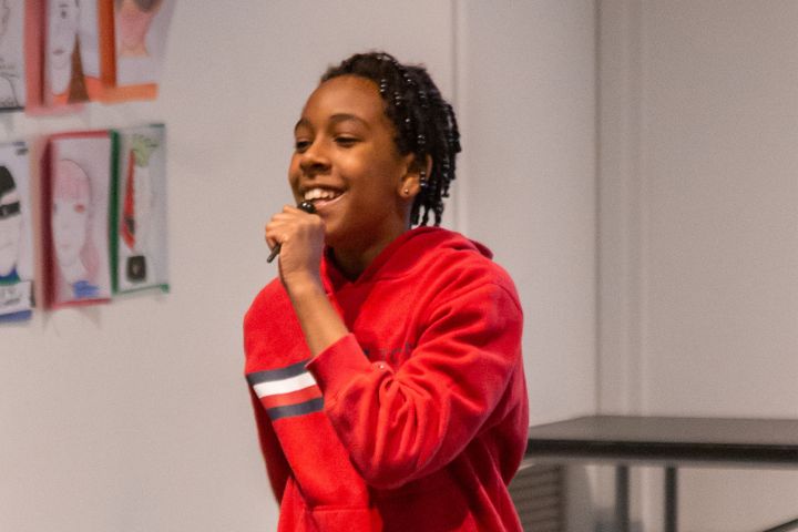 A young person in a red hoodie holds a microphone and smiles while speaking or singing indoors; artwork is visible on the wall behind them.