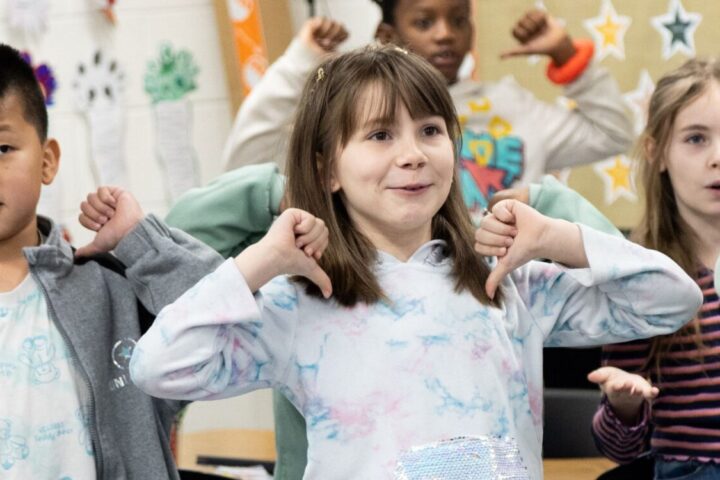 A group of young children stand indoors, smiling and raising their arms as if following along with a classroom activity. Colorful drawings are visible in the background.