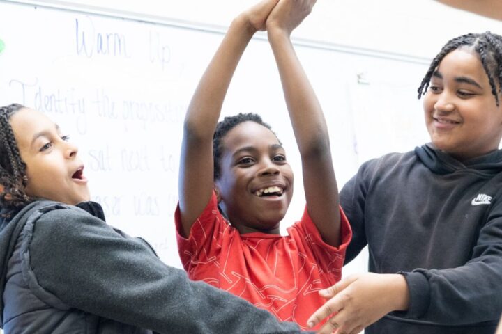 Three children stand together in a classroom, smiling and holding up their arms as if celebrating or playing a game. A whiteboard with writing is visible in the background.
