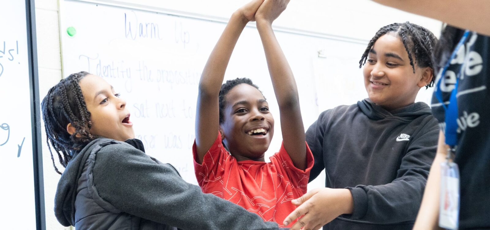 Three children stand together in a classroom, smiling and holding up their arms as if celebrating or playing a game. A whiteboard with writing is visible in the background.