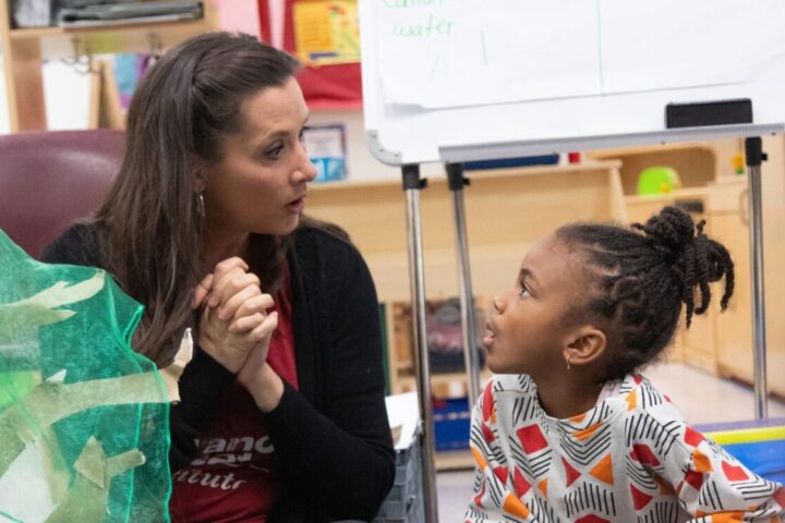 A woman and a young child sit facing each other in a classroom, engaged in conversation near a whiteboard and colorful materials.