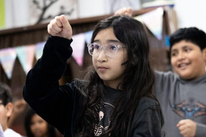 A girl with glasses raises her fist while a boy in the background smiles, both appearing engaged in a classroom setting.