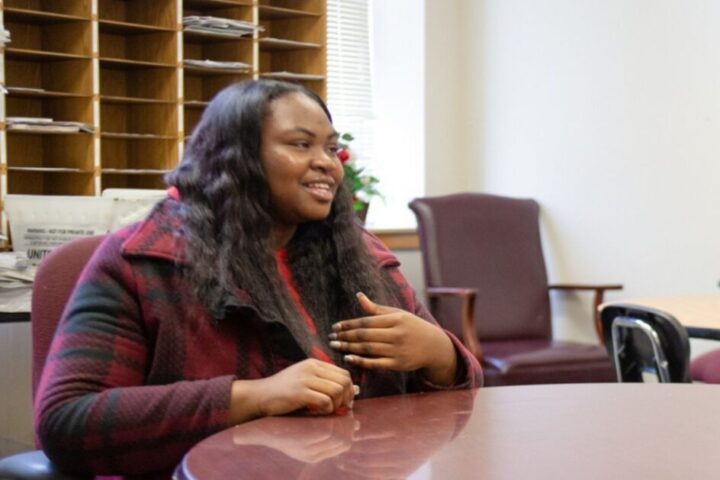 Two women sit across from each other at a round table in an office, engaged in conversation. One is holding a notepad and pen. Shelves and chairs are in the background.