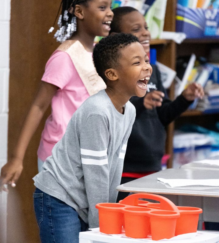 A young boy in a gray shirt laughs while playing a game with other children in a classroom setting.
