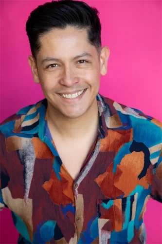 A man with short dark hair smiles at the camera, wearing a colorful patterned shirt, standing in front of a bright pink background.