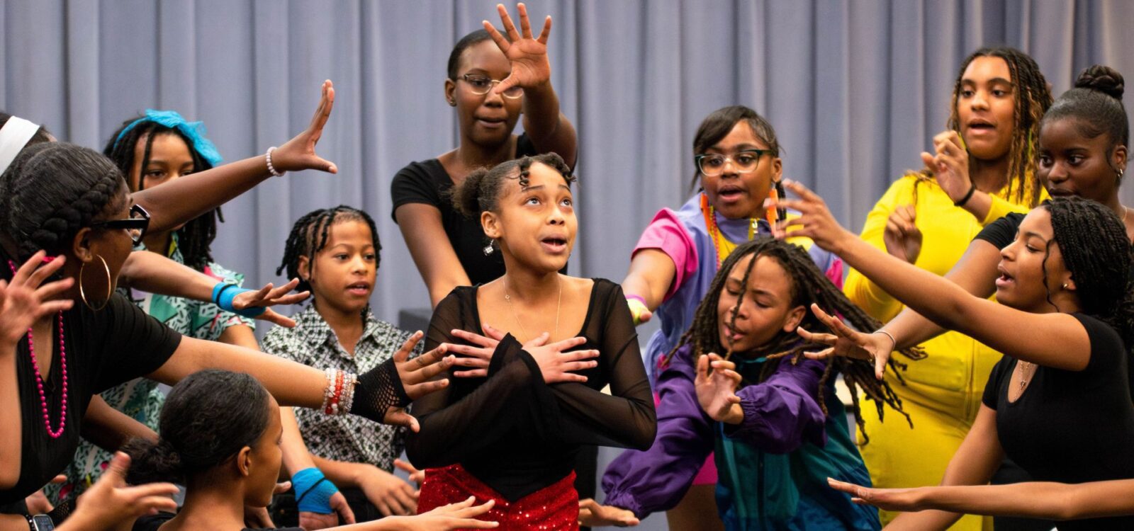 A group of young girls, some with hands raised, surround and focus on a girl in the center with her hands crossed on her chest, in front of a gray curtain.
