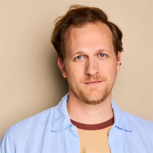 A man with light brown hair and a short beard looks at the camera, wearing a light blue shirt over a beige t-shirt, standing against a beige background.