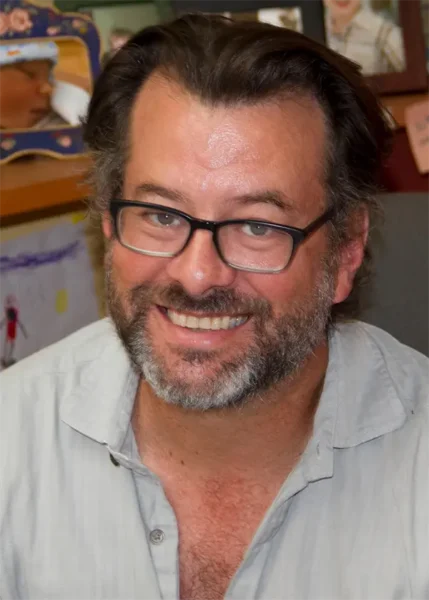 A man with dark hair, a beard, and glasses smiles at the camera. He is wearing a light grey shirt, and there are colorful drawings and photos in the background.