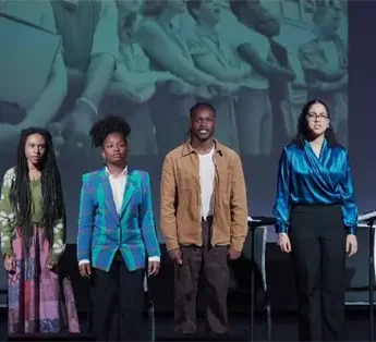 Four people stand on stage in front of chairs and a large projected black-and-white photo of people holding documents.
