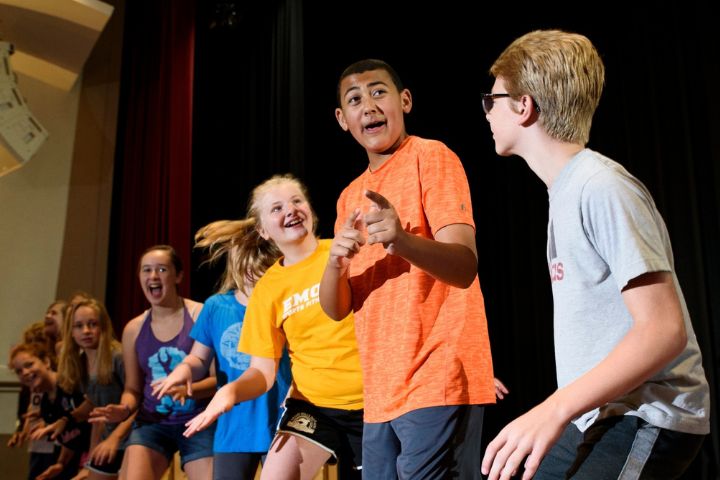 A group of young people rehearse on a stage, with one boy in an orange shirt gesturing and others smiling and watching him.