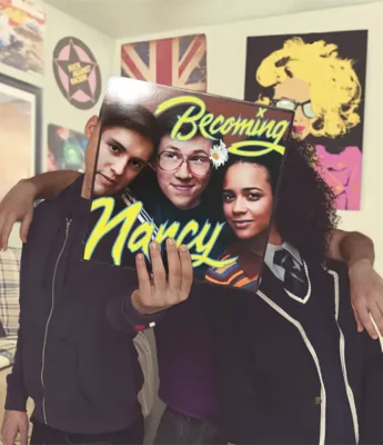 Three people pose in a room with one person holding a "Becoming Nancy" record in front of their faces, blending the cover art with their own. Posters decorate the wall in the background.