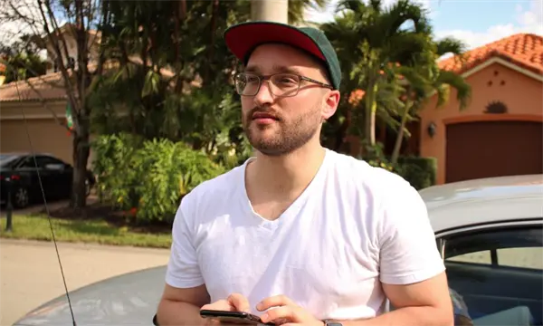 A man wearing glasses, a green cap, and a white t-shirt stands outdoors in front of a car and houses, holding a smartphone.