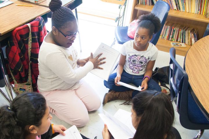 Four girls sit in a classroom in a small group, holding papers and engaging in discussion near bookshelves and desks.
