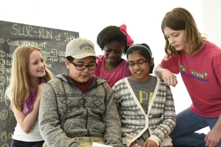 Five children are gathered together in a classroom, reading from a notebook. A chalkboard with writing is visible in the background.