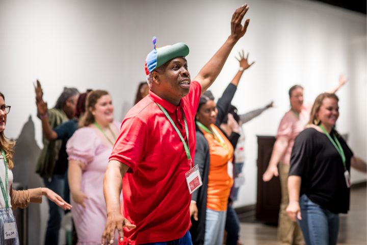 A group of people, some wearing name tags, stand indoors with arms raised, appearing to participate in an interactive group activity.