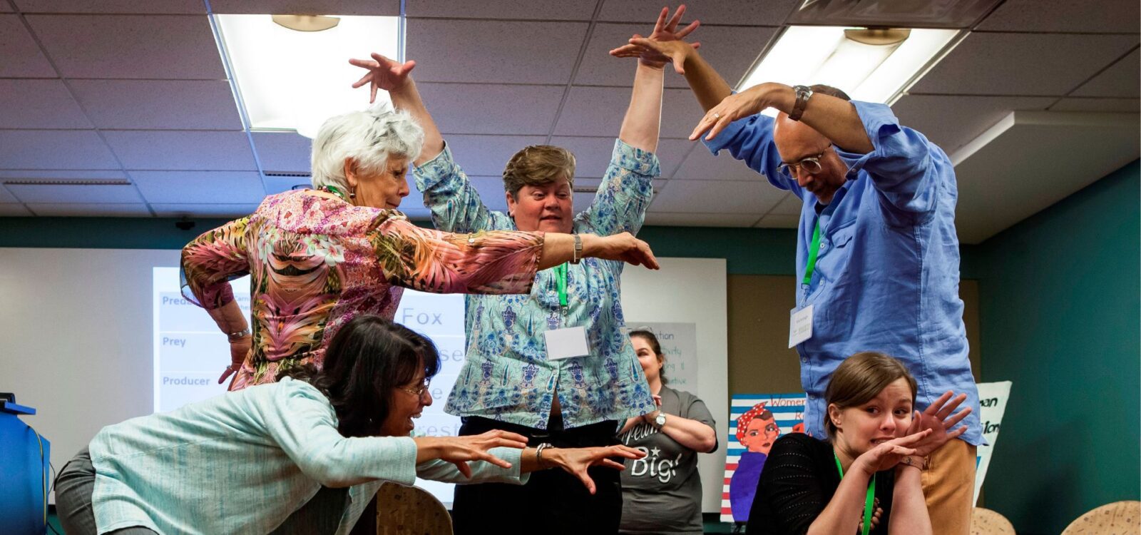 Six adults in a classroom stand and crouch in playful, dramatic poses with their arms raised and extended, appearing to participate in an interactive group activity.