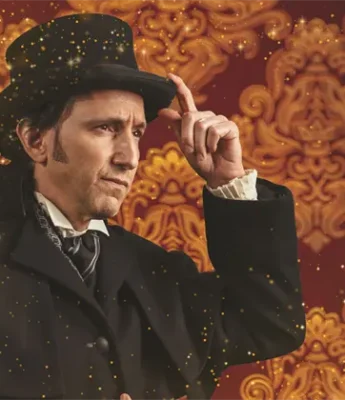 A man in a black coat and top hat poses against an ornate, gold-patterned red background with sparkling light effects.