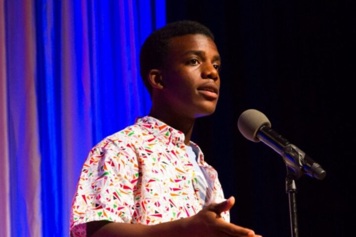 A young man in a patterned shirt speaks into a microphone on stage with blue and white curtains in the background.
