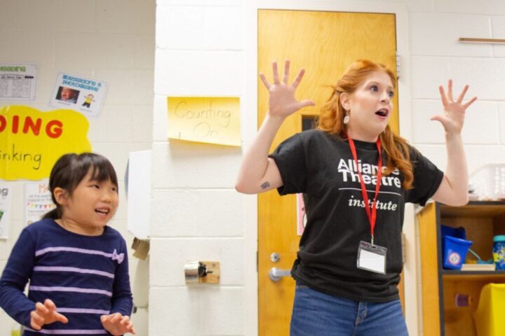 A teacher and two young students stand in a classroom with their hands raised and fingers spread, appearing to act out or express excitement.