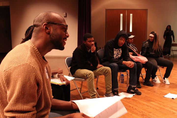 A group of people sit in a semi-circle on chairs in a room, some holding papers, as a man in the foreground speaks or leads a discussion.