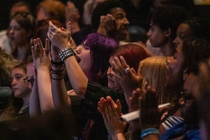 A diverse group of people seated closely together applaud enthusiastically at an indoor event.