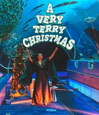A person in a festive outfit stands in an aquarium tunnel beneath a sign reading "A Very Terry Christmas," surrounded by fish and Christmas decorations.