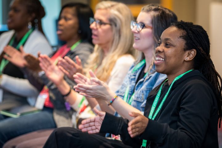 A group of people seated in a row, wearing green lanyards, clapping and smiling during an indoor event.