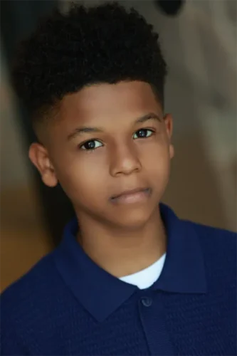 A young boy with short curly hair wearing a blue collared shirt looks directly at the camera with a neutral expression.