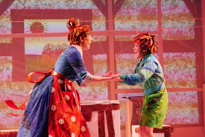 Two actors in colorful, leaf-themed costumes perform on stage in front of a painted backdrop featuring a sun and fields.