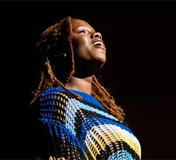 A person with long hair sings or speaks on stage under a spotlight, wearing a colorful, patterned outfit against a dark background.