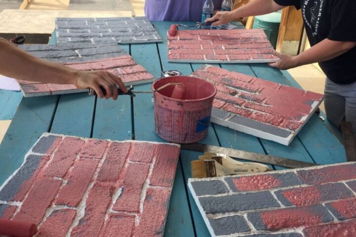Three people paint faux brick panels with red and gray paint using rollers on a blue wooden table; painting supplies are scattered on the table.