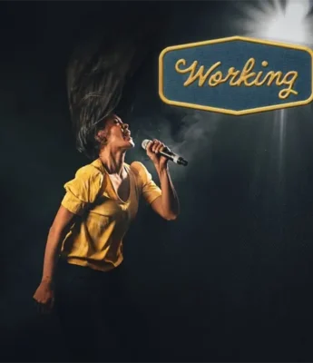 A person in a yellow shirt sings passionately into a microphone under a spotlight, with a sign reading "Working" illuminated above them.