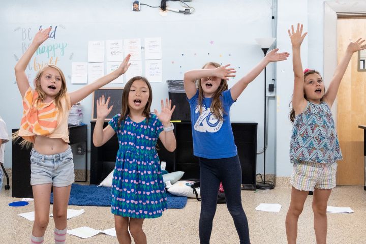 Four young girls stand indoors with their arms raised, appearing to sing or perform together. Papers are scattered on the floor behind them.