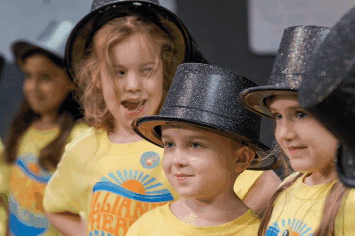 A group of young children wearing yellow shirts and black top hats stand together, smiling and looking in different directions.