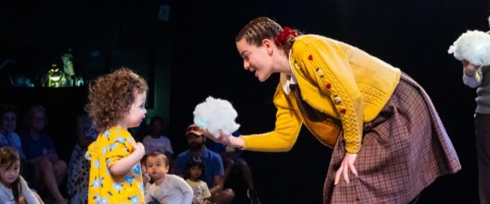 A woman in a yellow sweater and plaid skirt shows a fluffy white object to a young girl in a yellow dress on a stage as people watch in the background.