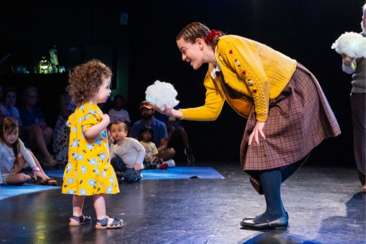 A woman in a yellow sweater and plaid skirt shows a fluffy white object to a young girl in a yellow dress on a stage as people watch in the background.