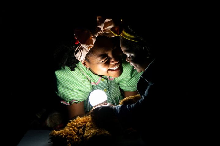 An adult and a child smile at each other in the dark, illuminated by a glowing light bulb. The adult wears a pink bow and green checkered shirt; the child holds a stuffed animal.