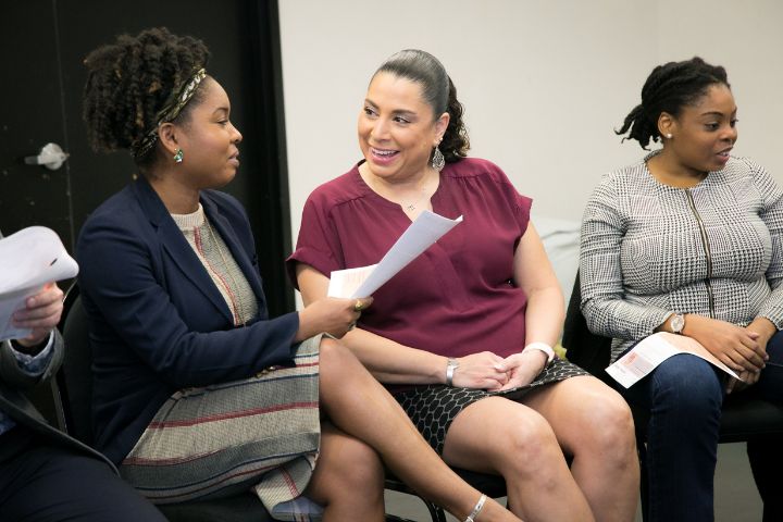 Three women sit in chairs indoors, holding papers and engaged in conversation, with one woman in the center smiling at another.