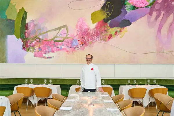 A chef stands in a modern restaurant dining room with wooden chairs, set tables, and a large colorful abstract mural on the wall behind him.