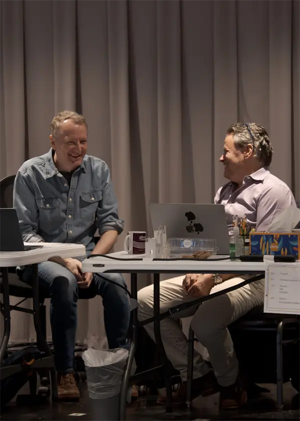 Two men sit at a table with laptops and papers, engaged in conversation and smiling, in front of a gray curtain backdrop.