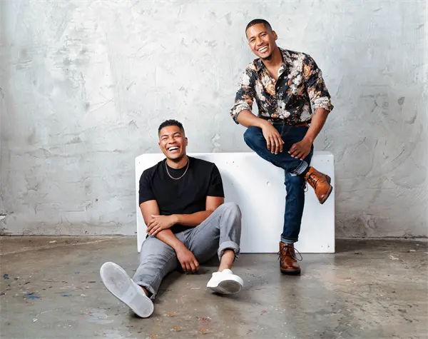 Two men pose in front of a textured gray wall; one sits on the floor in casual clothes while the other sits on a bench in a floral shirt and dress shoes, both smiling.