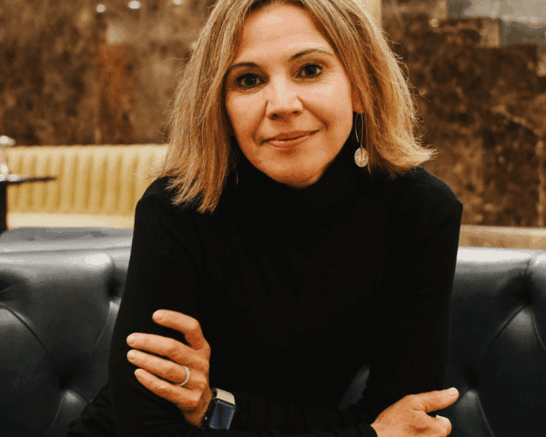 A woman with shoulder-length blonde hair, wearing a black turtleneck and earrings, sits on a blue sofa with her arms crossed in a warmly lit indoor setting.